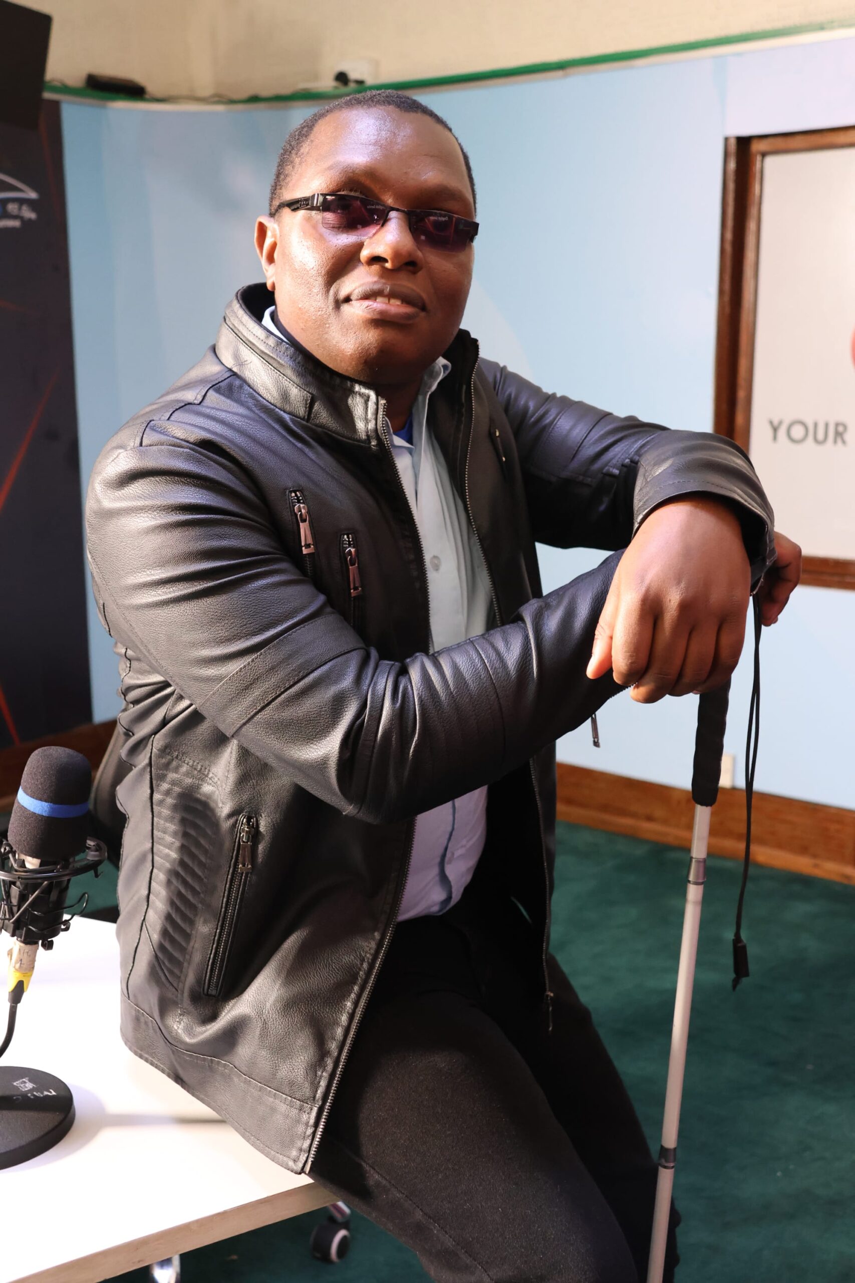Founder of Vuka Studios, a visually impaired voice-over artist, C.G Moses, seated in a recording studio wearing sunglasses and a black leather jacket, holding a white cane with a studio microphone beside him.